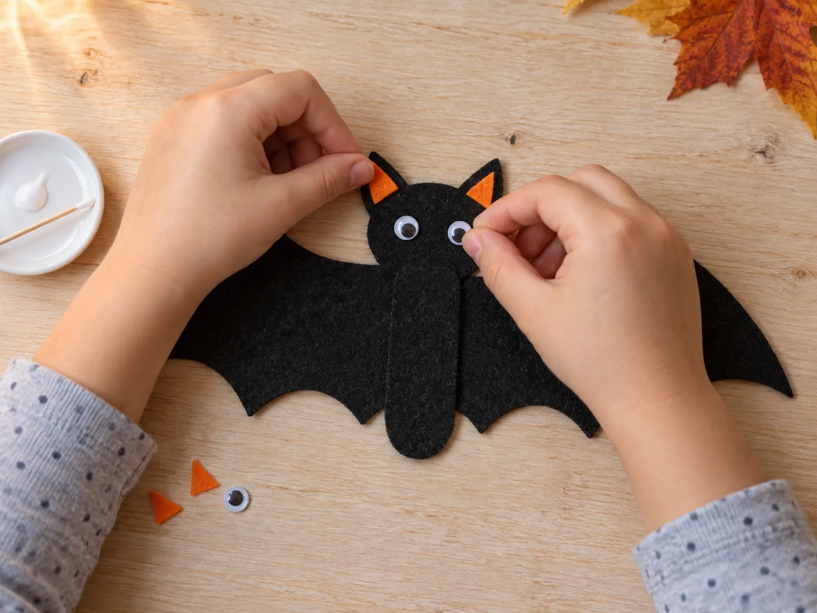 KI-generiertes Bild: Kinderhände kleben orange Ohren und kleine Wackelaugen auf eine schwarze Filz-Fledermaus