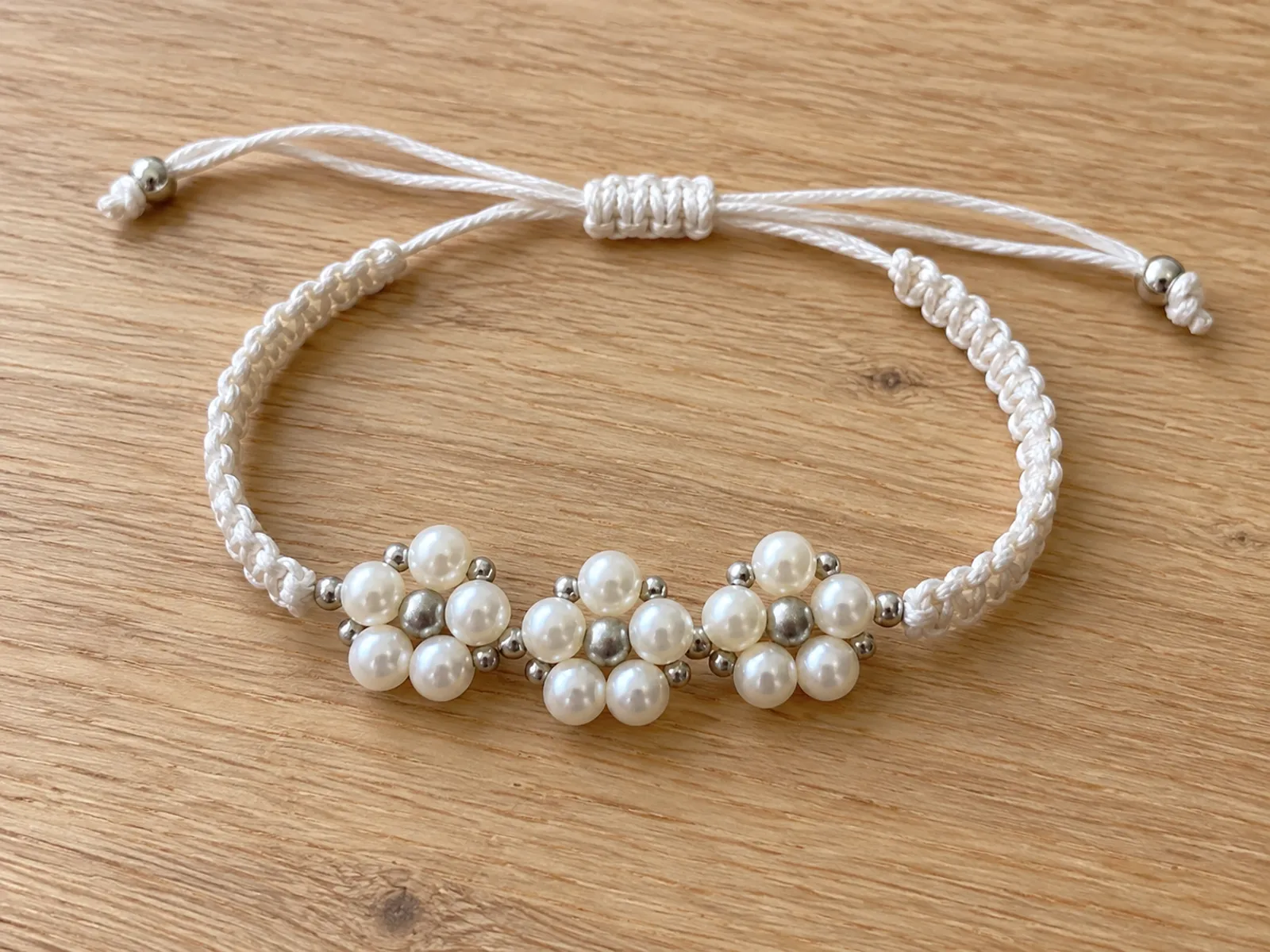 KI-generiertes Bild: fertiges weißes Makramee-Armband mit weißen Perlenblüten und mattsilbernen Metallperlen auf hellem Holztisch